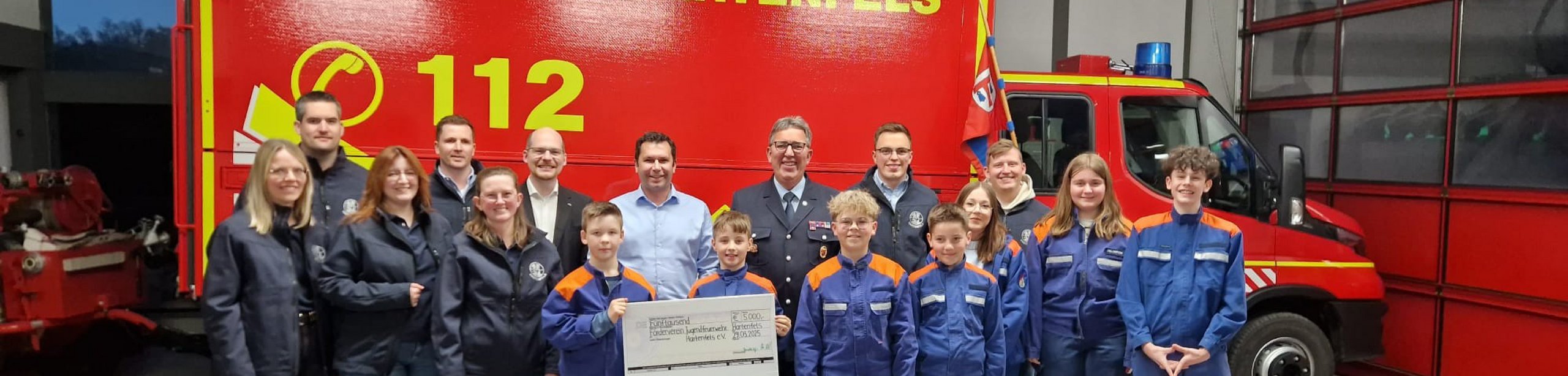 Feuerwehrgruppe mit großer Spende vor Einsatzfahrzeug Gruppenfoto mit Feuerwehrteam und Scheck vor rotem Fahrzeug