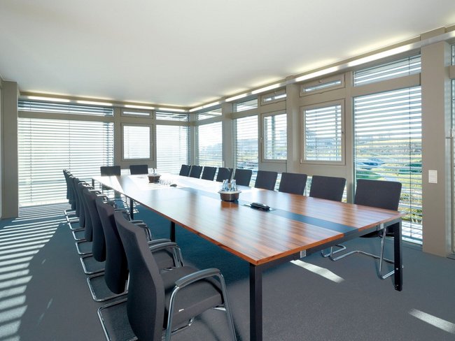Heller Konferenzraum mit großem Tisch und Stühlen, Fenster mit Jalousien