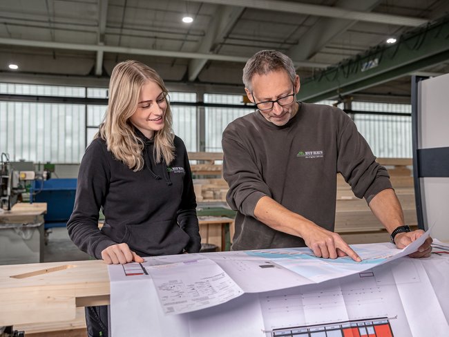 Zwei Personen in einer Werkstatt betrachten Pläne Ein Mann und eine Frau analysieren Baupläne in einer Holzwerkstatt.