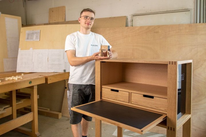 Mann hält Holzstück vor einem handgefertigten Schreibtisch mit Schubladen