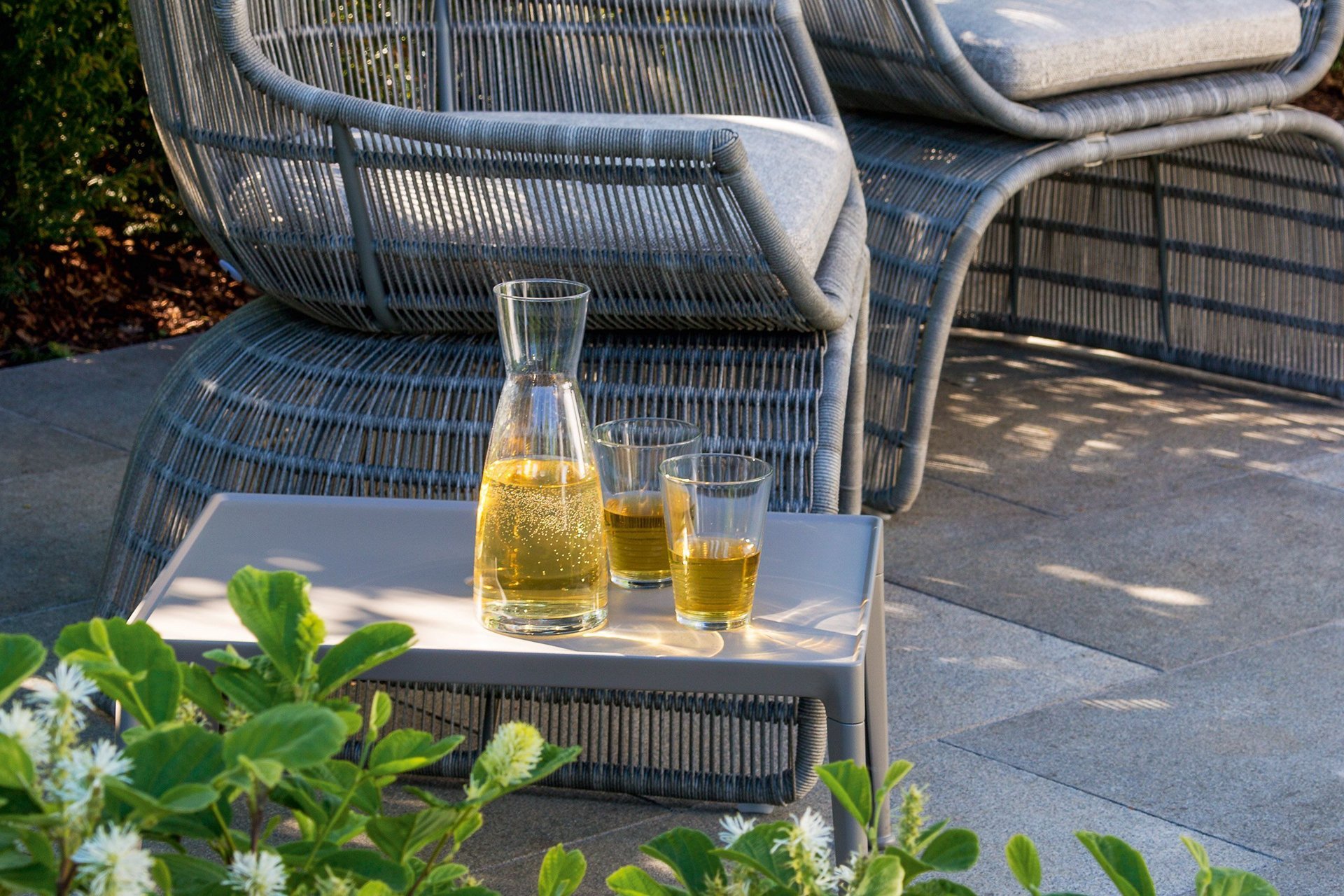 Glaswaren und Gartensessel bei Tageslicht Ein Wasserkaraffe und Gläser auf einem Tisch neben einem bequemen Sessel im Freien.