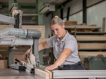 Frau arbeitet an einer Holzsäge in einer Werkstatt Frau in grauem Poloshirt bedient eine Holzsäge in einer Werkstatt