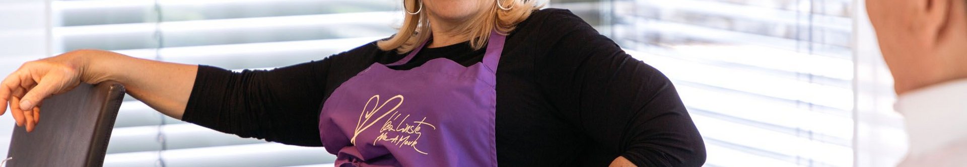 Lächelnde Frau mit lila Schürze am Tisch Frau in lila Schürze sitzt an einem Tisch und lächelt.