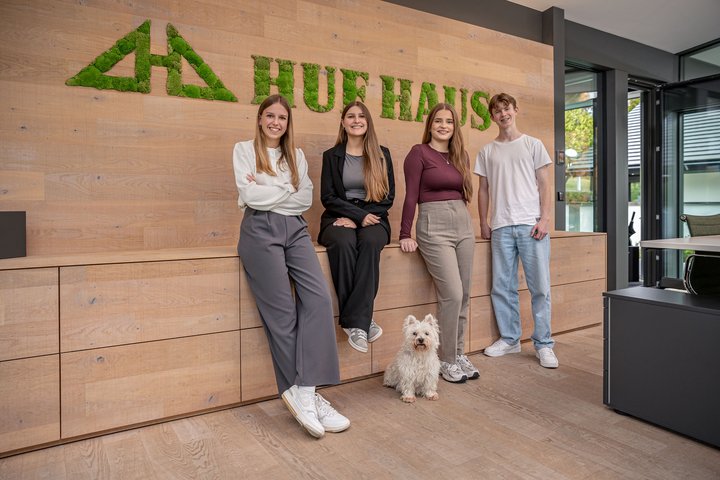 Teamfoto im Büro von HUF HAUS mit Hund Vier Personen posieren vor einem modernen Büro mit Pflanzenlogo