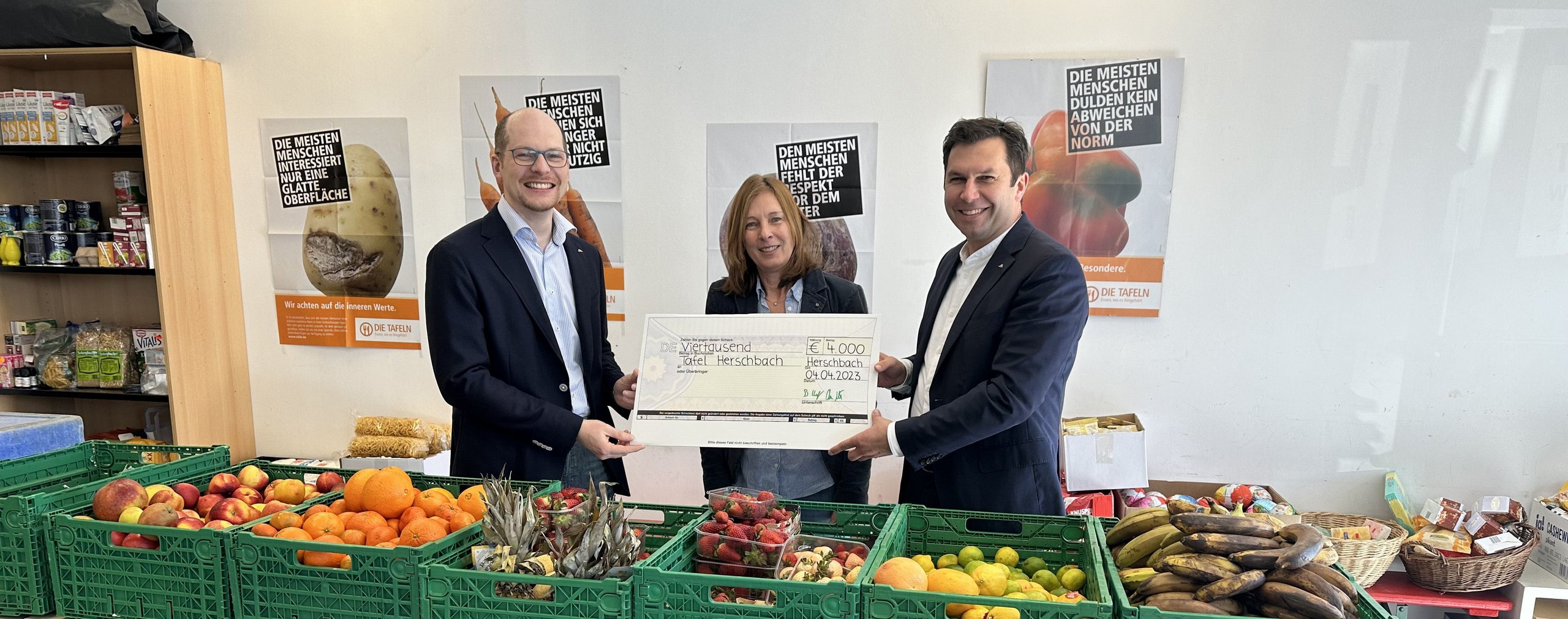 Spendenübergabe in einem Lebensmittelgeschäft Drei Personen übergeben einen großen Scheck vor Obst und Gemüse