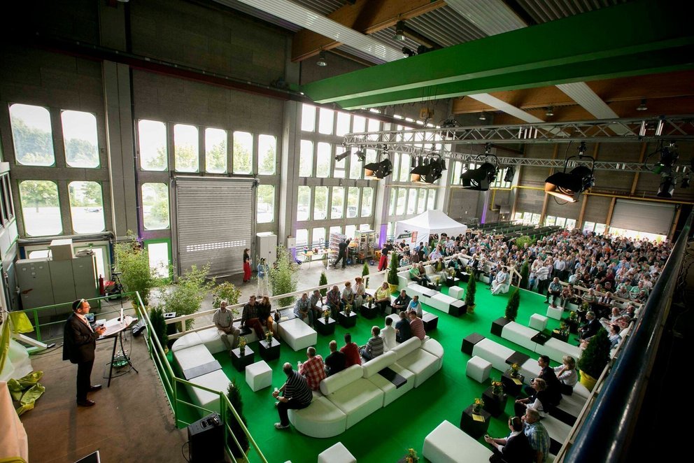 Konferenzveranstaltung in großer Halle mit Teilnehmern Teilnehmer sitzen auf weißen Bänken in einer modernen Halle bei einer Konferenz
