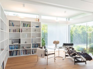 Helles Wohnzimmer mit großem Fenster, Bücherregal und relaxender Liege