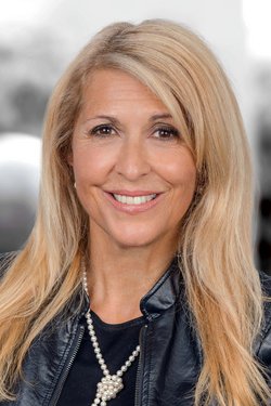 Frau mit blonden Haaren, Lederjacke und Schmuck lächelt freundlich.