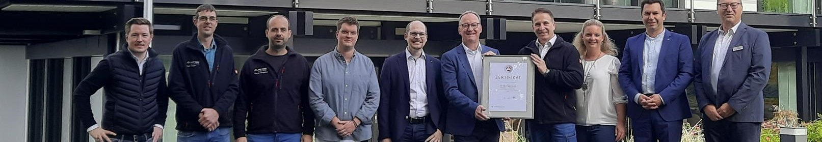 Gruppenfoto vor modernem Gebäude mit Zertifikat Menschen stehen zusammen vor einem modernen Haus mit einem Zertifikat
