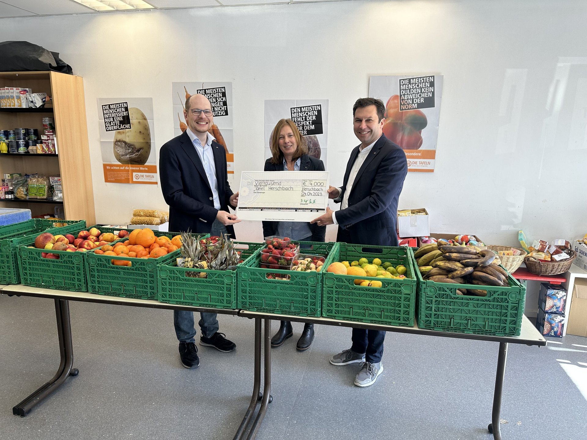 Spendenübergabe für lokale Lebensmittelausgabe Drei Personen übergeben einen großen Scheck in einem Raum mit Obst und Gemüse