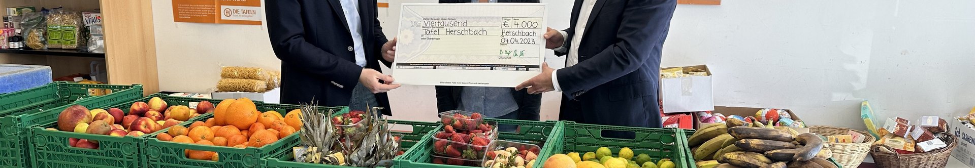 Spendenübergabe für lokale Lebensmittelausgabe Drei Personen übergeben einen großen Scheck in einem Raum mit Obst und Gemüse